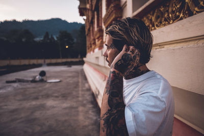 Man with tattoo looking away while standing by wall outdoors