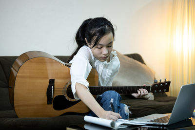 Cute girl writing in notebook while holding guitar guitar at home