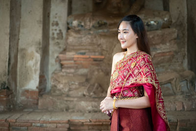 Thoughtful woman smiling while standing in traditional clothing against wall