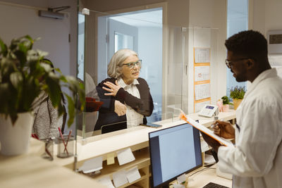 Senior female patient explaining health issues to male receptionist through transparent shield in clinic