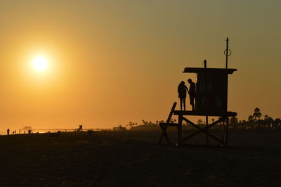 Silhouette lifeguards on hut at beach against orange sky