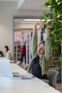 Tired woman adult learner stretching arms with closed eyes while studying in cozy library