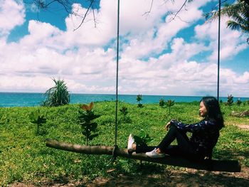 Side view of woman sitting on wooden swing against sky