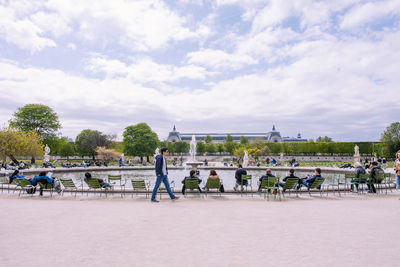 People relaxing on chair by fountain in park against cloudy sky