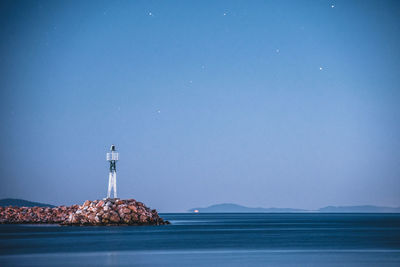 Lighthouse by sea against clear blue sky