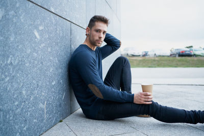 Side view of modern male in casual wear sitting near stone wall and br