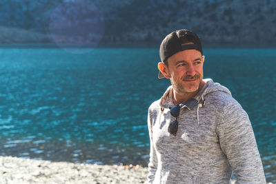 Attractive man standing in front of mountain lake with ball cap on looking away in grey hoodie.