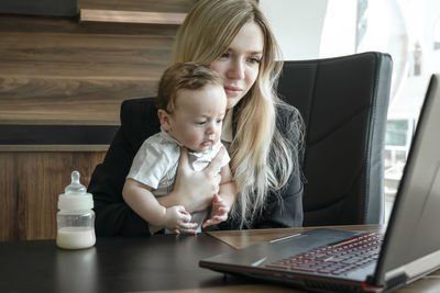 Young businesswoman holds small child in arms and communicates via video link via laptop in office.