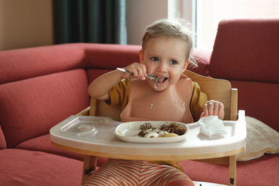 Toddler girl eating happily in high chair food by herself in the room. child having meal. happy kid