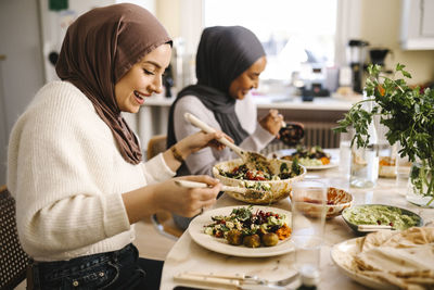 Smiling woman wearing hijab serving food on plate sitting at dining table