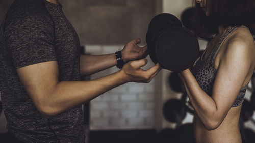 Midsection of instructor helping woman during lifting dumbbells in gym