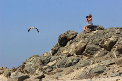 Low angle view of woman looking through binoculars on cliff against clear sky