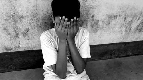Boy covering face with hands sitting by wall