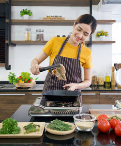 Young asian woman dressed in an apron use tongs to tong the steak from the hot pan.