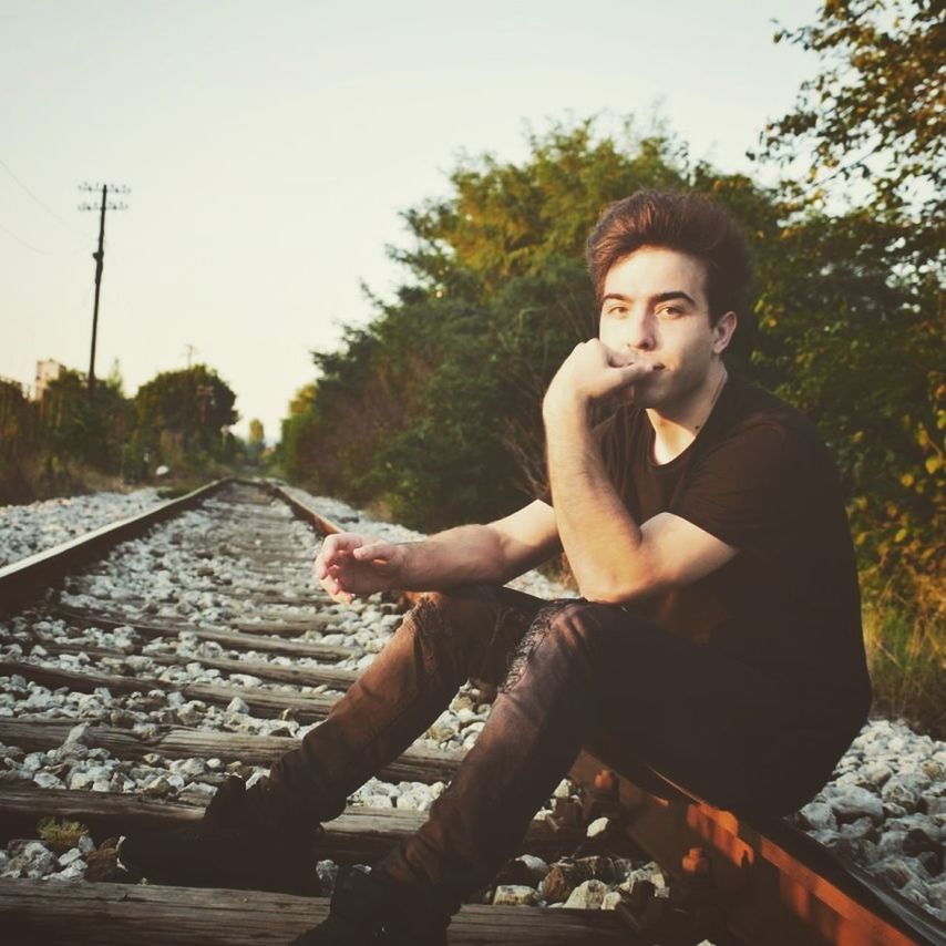 one person, young adult, sitting, young men, leisure activity, railroad track, track, lifestyles, real people, casual clothing, nature, rail transportation, portrait, sky, looking at camera, plant, full length, day, outdoors, contemplation, hairstyle