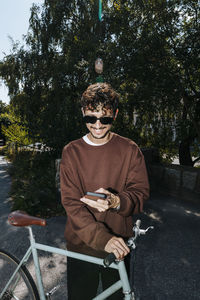 Smiling young man using smart phone while standing with bicycle at park