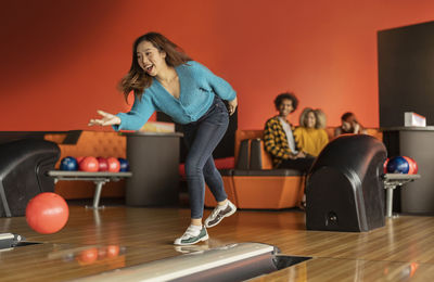 Young woman throwing ball with friends sitting in background at bowling alley