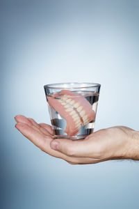Cropped hands of person holding dentures in glass of water against blue background