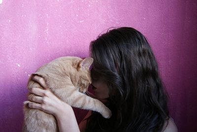 Close-up of young woman kissing cat against pink wall