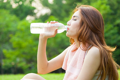 Woman drinking water from bottle against trees