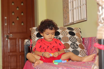 An indian little child is playing with toys sitting on the sofa inside the house