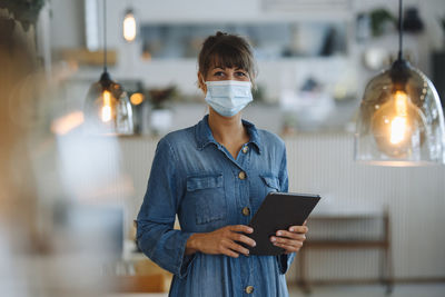 Female owner wearing protective face mask using digital tablet while standing in cafe