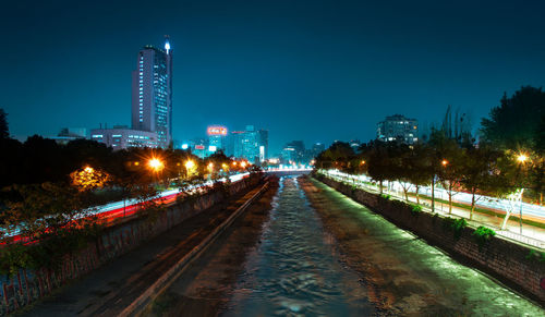 Illuminated street by buildings against sky at night