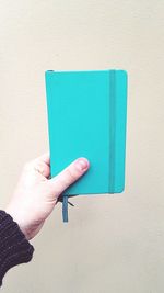 Cropped hand holding blue diary against wall