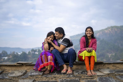 Full length of mother and girl sitting on rock against sky