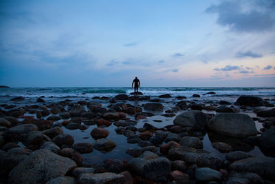 Silhouette person standing at rocky beach during dusk