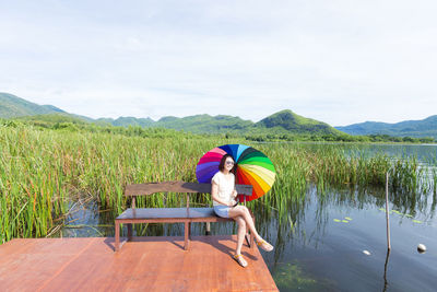 Young woman sitting on bench at jetty over lake against sky