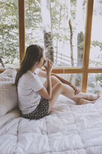 Young woman sitting on bed, drinking coffee and looking through window