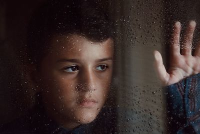 Close-up of thoughtful boy looking through wet glass window