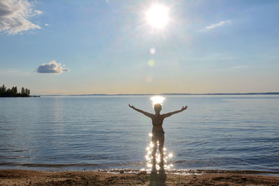 Rear view of woman with arms outstretched standing at beach against sky on sunny day
