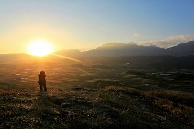 Rear view of person standing on field against mountains and sky during sunset