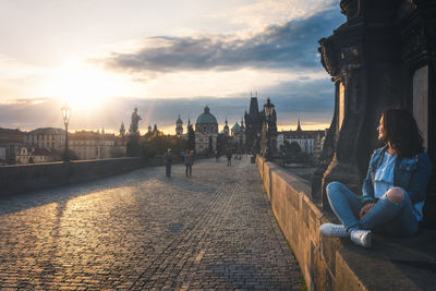 Woman sitting at charles bridge against sky during sunset