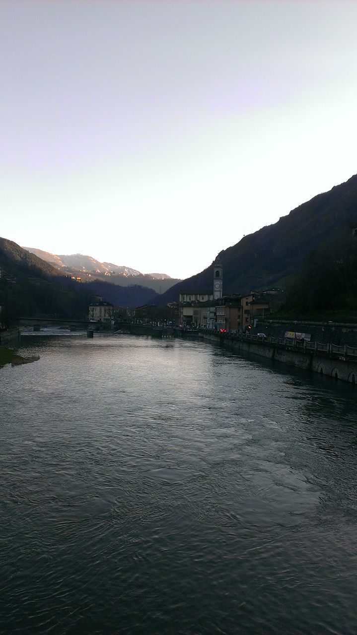 water, mountain, clear sky, copy space, tranquil scene, waterfront, tranquility, built structure, river, mountain range, scenics, architecture, beauty in nature, nature, lake, building exterior, rippled, outdoors, sky, day