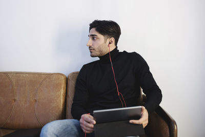 Young man looking away while listening music through digital tablet on sofa at office