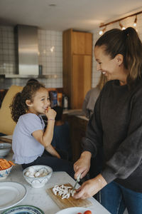 Smiling girl sitting near mother cutting mushroom while standing in kitchen at home