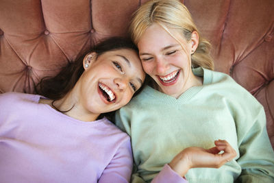 High angle of cheerful teenage sisters chilling on couch and laughing while having fun together at weekend