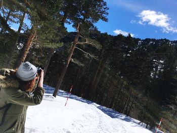Rear view of woman photographing on snow covered land against trees and sky