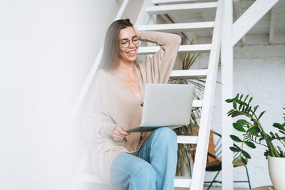 Young woman with long hair in cardigan working on laptop sitting on stairs at home