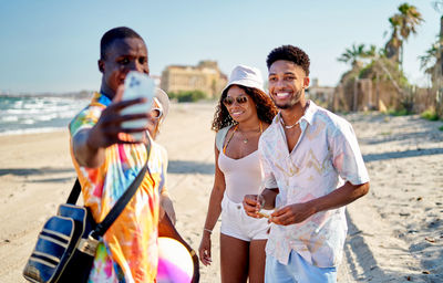 Black man using smartphone to take selfie with multiracial friends on sunny summer day on sandy beach