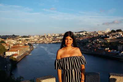 Portrait of young woman standing against river in city