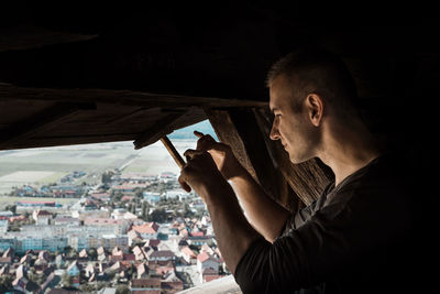 Side view of man looking through window at cityscape