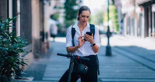 Woman using mobile phone while standing with push scooter in city