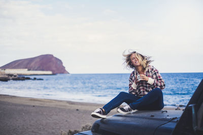 Portrait of cheerful young woman tossing hair on vehicle at beach against sky