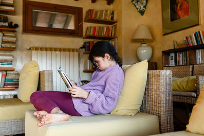 Side view of dark haired girl in lilac pullover reading a book in yellow armchair