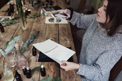 High angle view of mid adult female owner photographing brochure while sitting at table in workshop