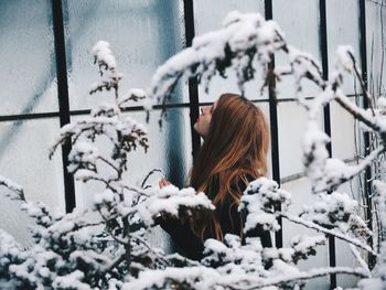 Side view of woman standing against window seen through branches during winter